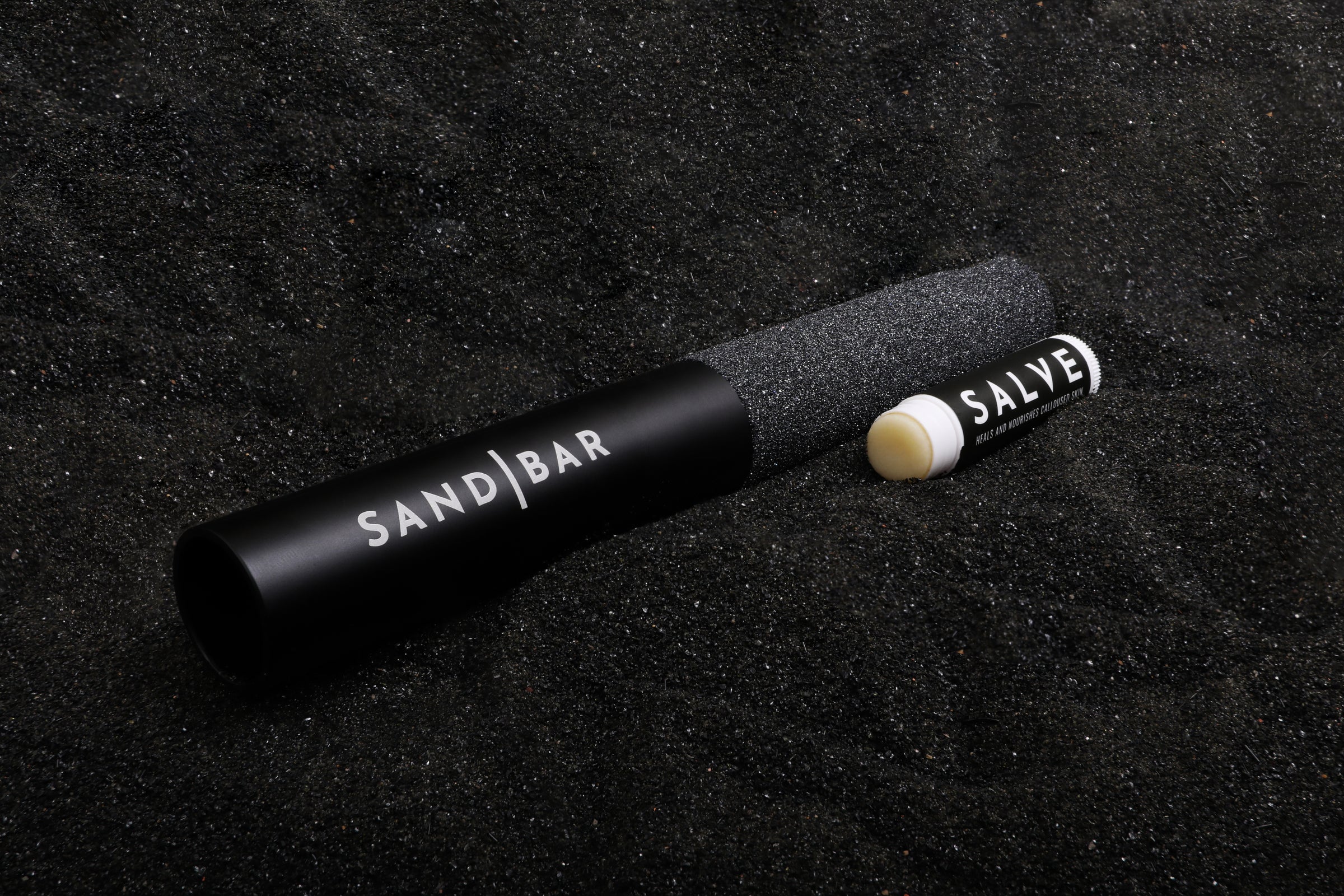 8" original SandBar callus file in a bed of black sand sitting next to the Sandbar Salve that comes in a ChapStick tube.
