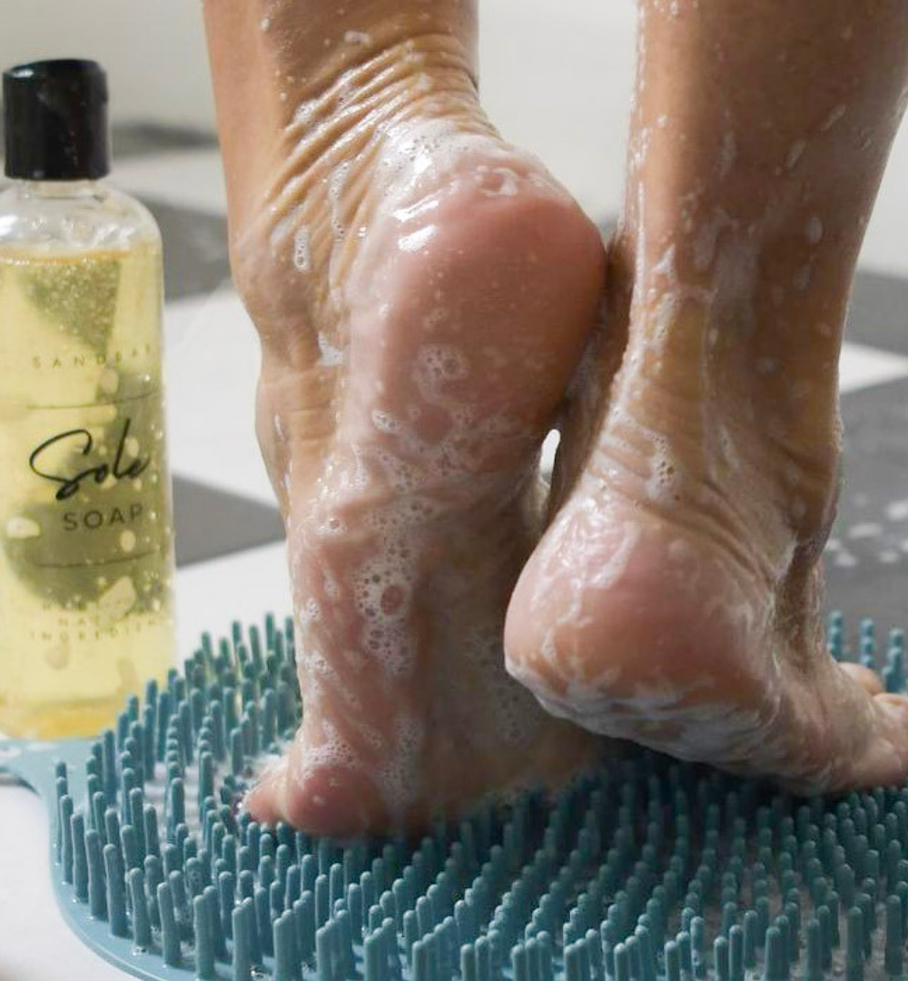 Feet in the shower all sudsy with bubbles standing on the foot scrubber mat next to the Sole Soap for perfectly clean feet.