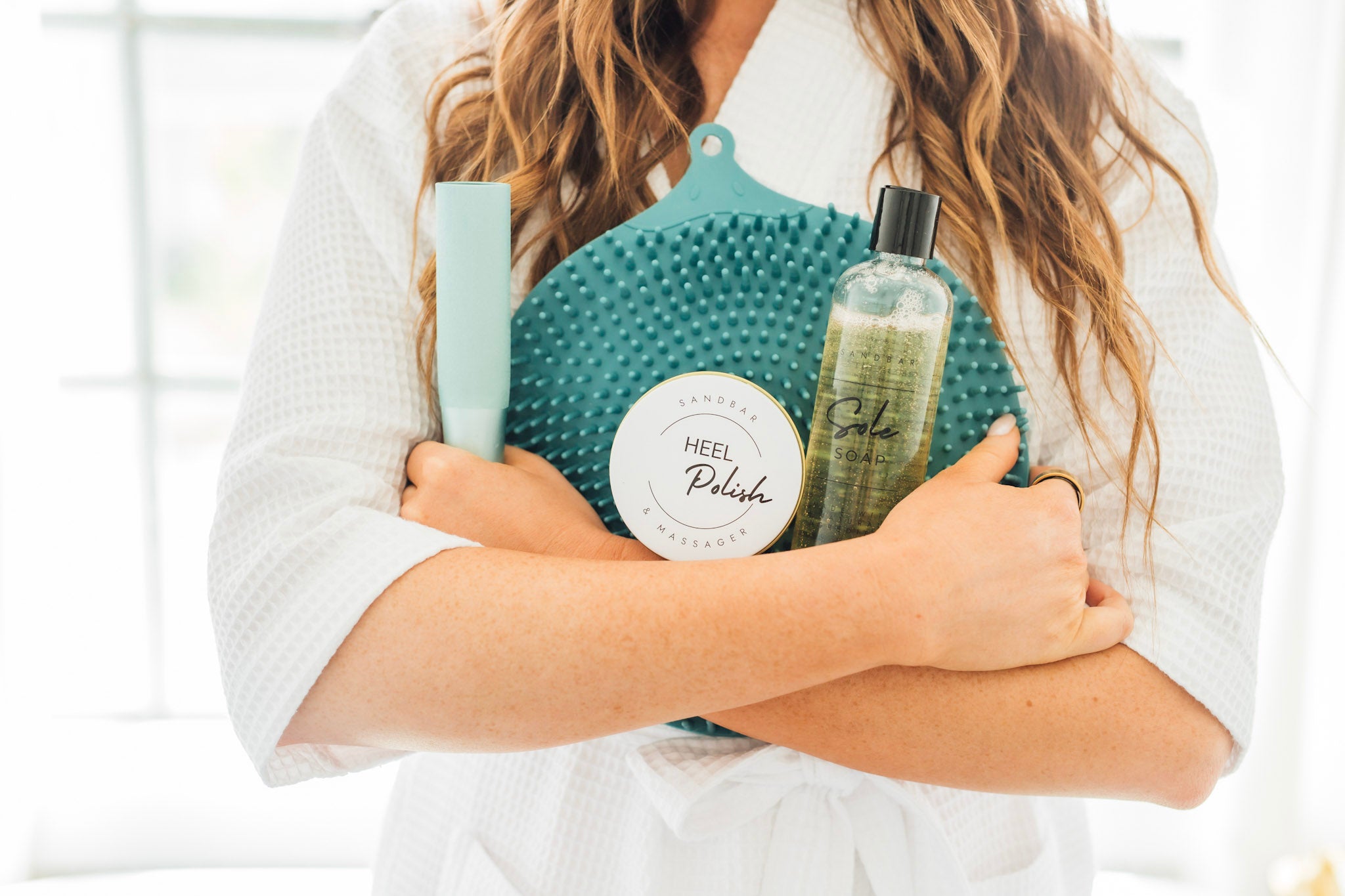 Women in a robe, holding/hugging her Heel Revival Regimen . Which includes the Foot Scrubber Mat, Sole Soap, Foot File and Heel Polish.
