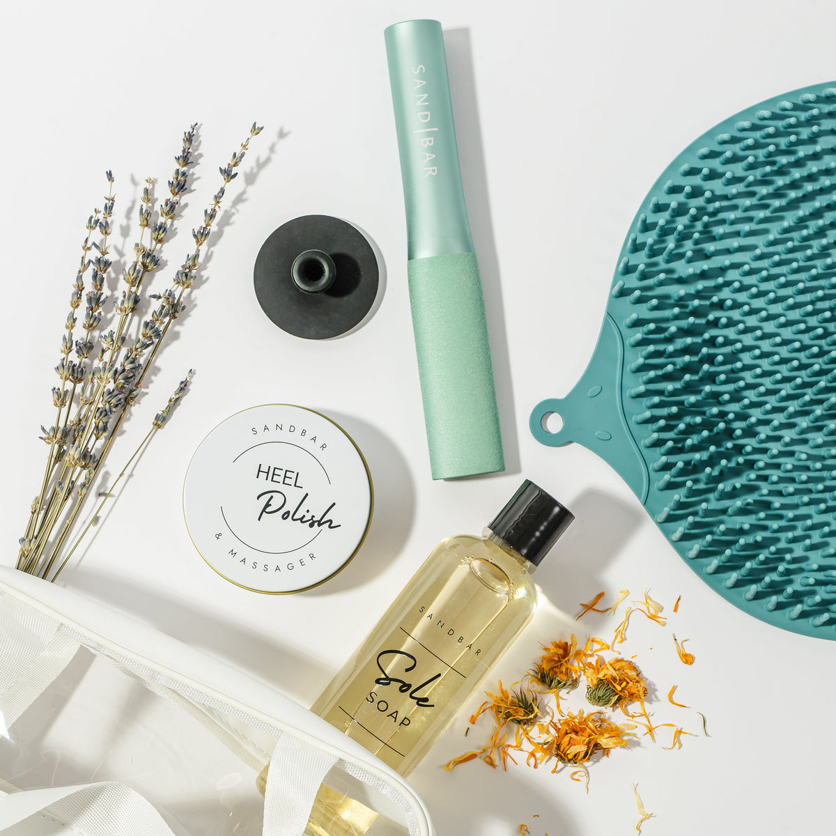 Heel Revival Regimen products laid out. Foot scrubber mat, Sole Soap, Foot File, Heel Polish and Applicator. It includes a white carrying case and also displays some natural ingredients that come in the sole soap and Heel Polish