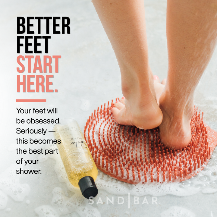 Close up of a woman's feet using an exfoliating shower mat and foot soap in the shower with feet lathered next to a bottle of sole soap.