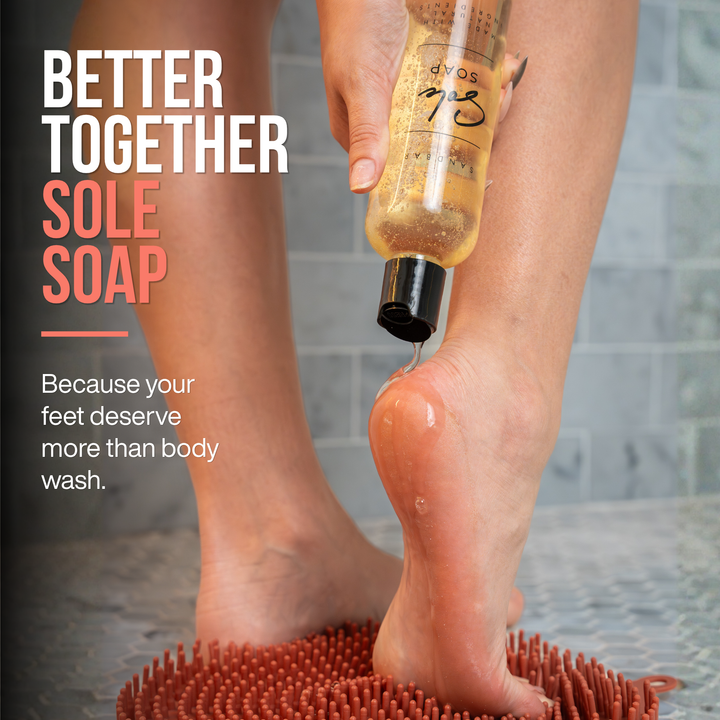 A woman pouring sole soap from a clear bottle with a black cap onto the heel of their foot, standing on a textured shower scrubber foot mat in a shower. Text on the left reads 'BETTER TOGETHER SOLE SOAP' and 'Because your feet deserve more than body wash.