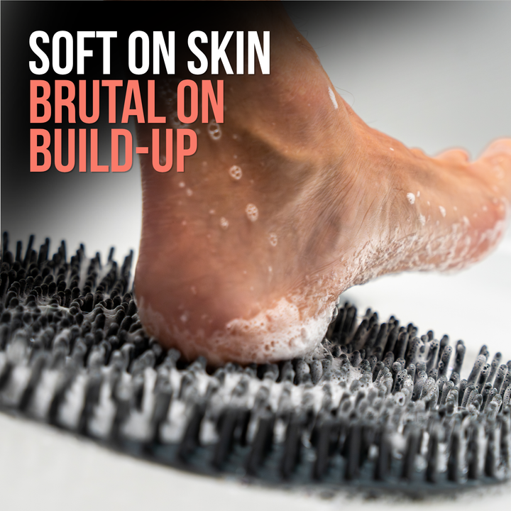 Close-up of a foot on a dark gray SandBar silicone scrubber mat showing flexible bristles exfoliating buildup on the heel while remaining gentle on skin, overlaid with the text 'SOFT ON SKIN BRUTAL ON BUILD-UP'. 