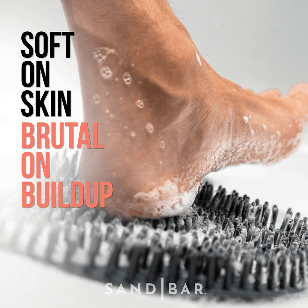 Close-up of a heel scrubbing across the SandBar shower foot scrubber mat with soap foam, gently exfoliating rough, built-up skin. Text reads “Soft on Skin, Brutal on Buildup.”